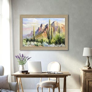 May include: A framed landscape oil painting depicts a desert scene with mountains and cacti. The artwork features a palette of greens, blues, and oranges, with a light-colored wooden frame. The painting hangs above a wooden desk with a laptop and chair.