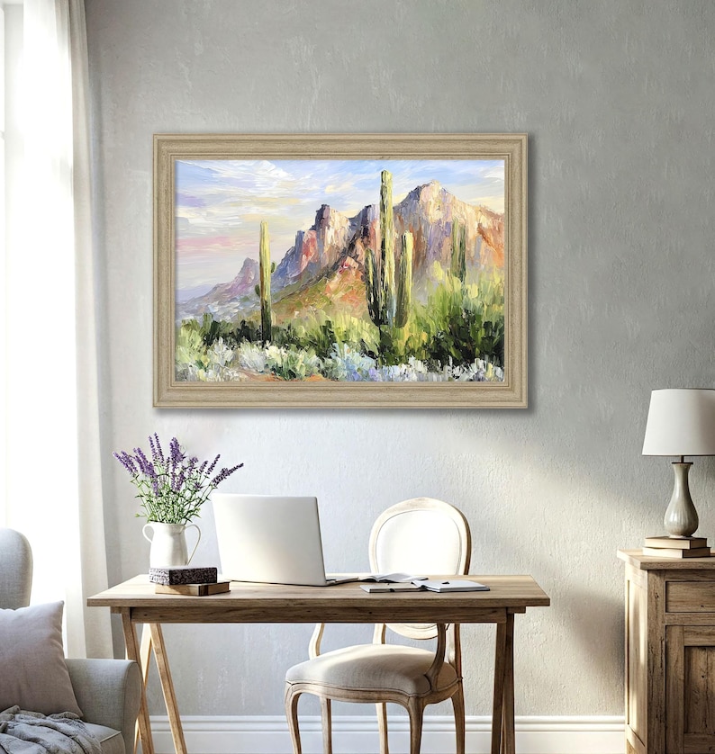 May include: A framed landscape oil painting depicts a desert scene with mountains and cacti. The artwork features a palette of greens, blues, and oranges, with a light-colored wooden frame. The painting hangs above a wooden desk with a laptop and chair.