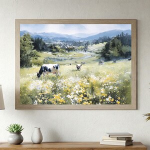 May include: A framed landscape painting depicts a serene pasture scene. Cows graze in a field of wildflowers, with rolling hills and mountains in the background. The artwork is encased in a light brown wooden frame.
