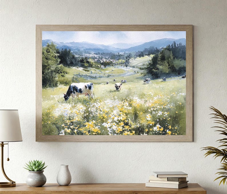 May include: A framed landscape painting depicts a serene pasture scene. Cows graze in a field of wildflowers, with rolling hills and mountains in the background. The artwork is encased in a light brown wooden frame.