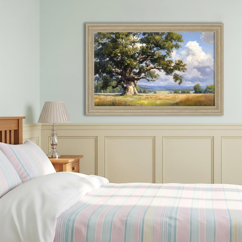May include: A framed landscape painting shows a large oak tree in a field under a blue sky with white clouds. The tree has green leaves, and the field is yellow and green. The frame is a light beige colour.