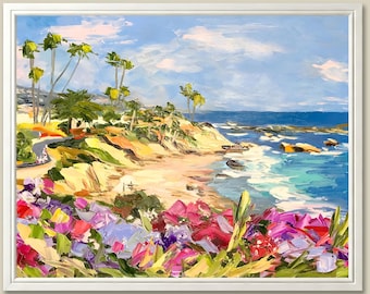 Op de afbeelding: Olieverfschilderij in een witte lijst dat een kustlandschap weergeeft. Het kunstwerk toont een strand met kleurrijke bloemen, een zandstrand en de zee onder een blauwe lucht met wolken. Palmbomen zijn zichtbaar op de kliffen.