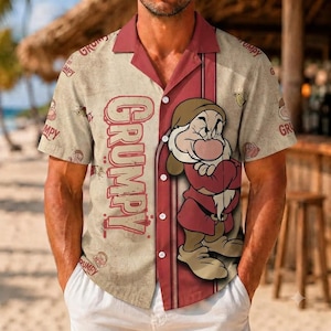 May include: A short-sleeved button-down shirt in a beige, distressed-look fabric featuring a large graphic of the character Grumpy from Snow White. The shirt includes the word GRUMPY in red text and a repeating pattern of small character icons.