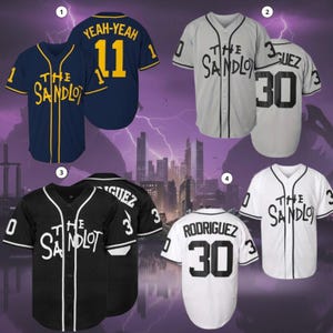 Puede incluir: Camisetas de béisbol en azul marino, gris, negro y blanco. Cada camiseta presenta el texto "The Sandlot" y el nombre y número de un jugador. Las camisetas tienen botones en la parte delantera y mangas cortas, con detalles de color contrastantes.