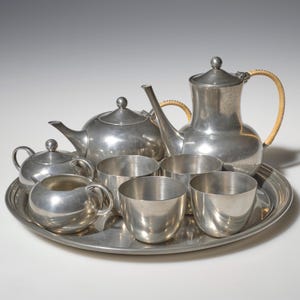 May include: A silver-toned metal tea set consisting of a tall teapot with a wrapped handle, a shorter teapot, a lidded sugar bowl, a creamer, and three cups, all arranged on a large round serving tray with a smooth, polished metallic finish.