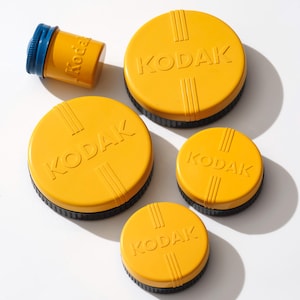 May include: A collection of vintage Kodak film canisters and containers. The items are yellow with black and blue accents. The Kodak logo is prominently displayed on the lids. The items vary in size, suggesting different film formats.
