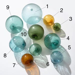 Pode incluir: Uma coleção de dez boias de pesca de vidro em vários tamanhos e cores, incluindo transparente, âmbar, verde e azul. As esferas estão dispostas sobre uma superfície branca, projetando sombras suaves.