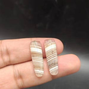 Puede incluir: Dos piedras preciosas rectangulares pulidas con un patrón a rayas de color blanco, marrón y beige. Las piedras se sostienen entre los dedos sobre un fondo oscuro. Las piedras preciosas tienen un aspecto translúcido.