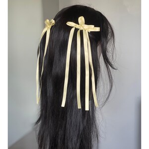 Könnte beinhalten: Schwarzes Haar, das mit zwei gelben Haaraccessoires aus Bändern gestylt wurde. Ein Accessoire ist eine kleine Schleife, das andere eine größere Schleife mit langen, herabhängenden Bändern. Die Bänder sind hellgelb.