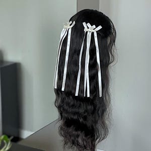 Puede incluir: Una peluca negra con cabello largo y ondulado, adornada con cintas de satén blancas atadas en lazos. La peluca se exhibe en un soporte negro contra una pared gris neutra, mostrando la textura del cabello y los detalles de las cintas.