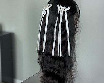 May include: A black wig with long, wavy hair is adorned with white satin ribbons. The ribbons are tied in bows at the top of the wig and cascade down the length of the hair, creating a decorative and elegant look.