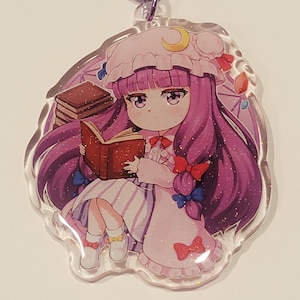 Könnte beinhalten: Ein klarer Acryl-Anhänger mit einer Cartoon-Figur mit langen lila Haaren, einem rosa Kleid und einem Buch. Der Anhänger hat einen lila Verschluss und ein glitzerndes Finish. Die Figur trägt einen Hut mit einem Halbmond.