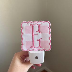 Może przedstawiać: Biała lampka nocna z różową haftowaną literą "F" na różowo-białym tle w kratkę. Lampka nocna ma falisty brzeg i białą podstawę.