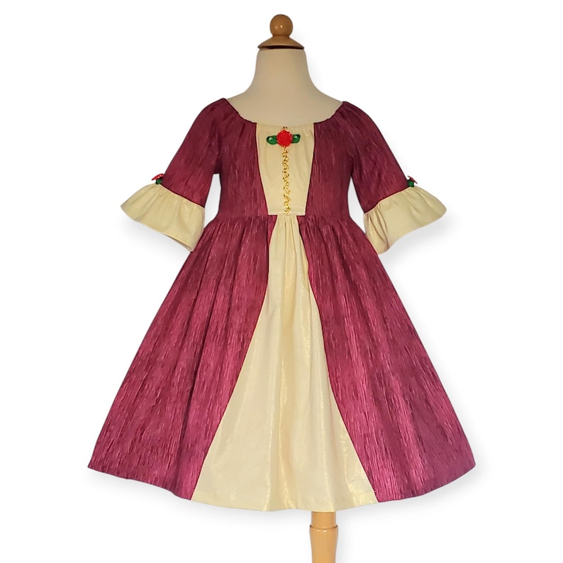 May include: A burgundy and cream-coloured dress with a scoop neckline and short sleeves. The dress features a cream-coloured panel down the front, adorned with gold detailing and a red rose accent. The sleeves have cream-coloured cuffs.