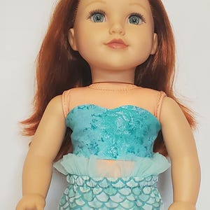 May include: A doll with red hair and blue eyes, wearing a mermaid-themed outfit. The outfit includes a teal top, a ruffled skirt, and a scale-patterned bottom. The doll has a fair complexion and is posed in a neutral position.