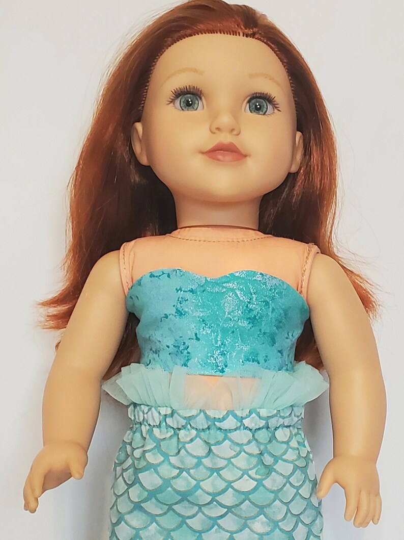 May include: A doll with red hair and blue eyes, wearing a mermaid-themed outfit. The outfit includes a teal top, a ruffled skirt, and a scale-patterned bottom. The doll has a fair complexion and is posed in a neutral position.