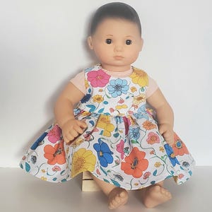 May include: A doll wearing a white dress with a colourful floral print. The dress has a pink bodice and a ruffled skirt. The doll has brown hair and brown eyes.