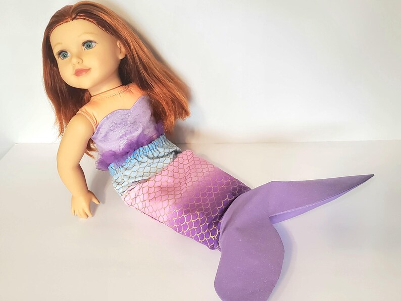 以下が含まれることがあります： 赤い髪と青い目の人形が人魚のコスチュームを着ています。コスチュームは紫色のトップス、ピンクと紫色の鱗模様のスカート、大きな紫色の尾が特徴です。人形は白い表面に置かれています。