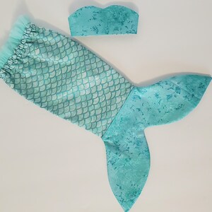 May include: A turquoise mermaid tail and matching top. The tail features a scale pattern on the upper portion and a solid, textured design on the fin. The top is a bandeau style, also in turquoise.