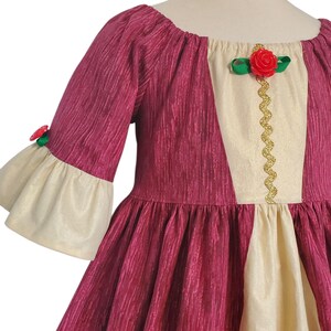 May include: A burgundy dress with cream-coloured accents. The dress features a scoop neckline, elbow-length sleeves with cream-coloured bell cuffs, and a full skirt. A cream-coloured panel is centered on the bodice, adorned with a gold trim and a red rose.