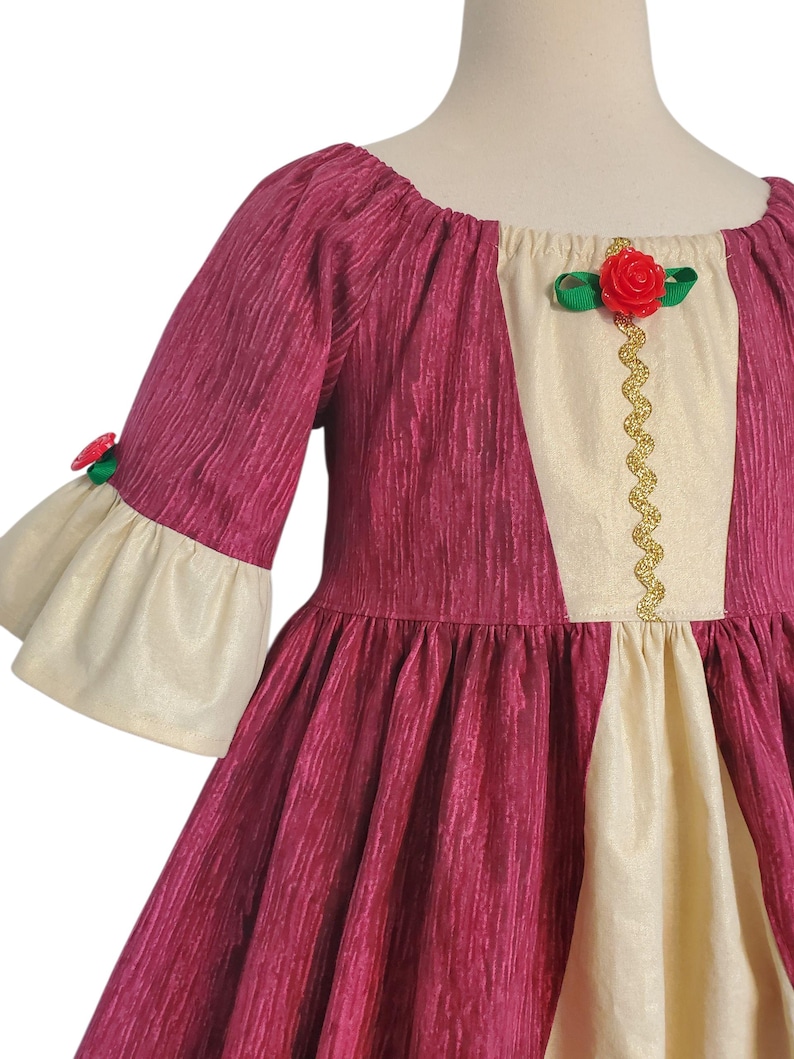 May include: A burgundy dress with cream-coloured accents. The dress features a scoop neckline, elbow-length sleeves with cream-coloured bell cuffs, and a full skirt. A cream-coloured panel is centered on the bodice, adorned with a gold trim and a red rose.