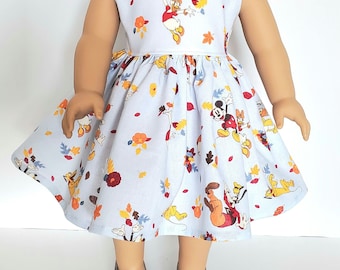 Puede incluir: Un vestido azul claro sin mangas con un estampado de Mickey Mouse, Minnie Mouse, Donald Duck y Goofy en hojas de otoño. El vestido tiene una falda fruncida y es perfecto para una muñeca.