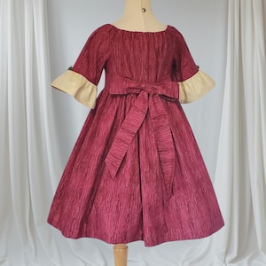 May include: A burgundy dress with textured fabric, featuring a boat neckline and elbow-length sleeves with cream-coloured cuffs. A matching bow adorns the waist. The dress is displayed on a mannequin.