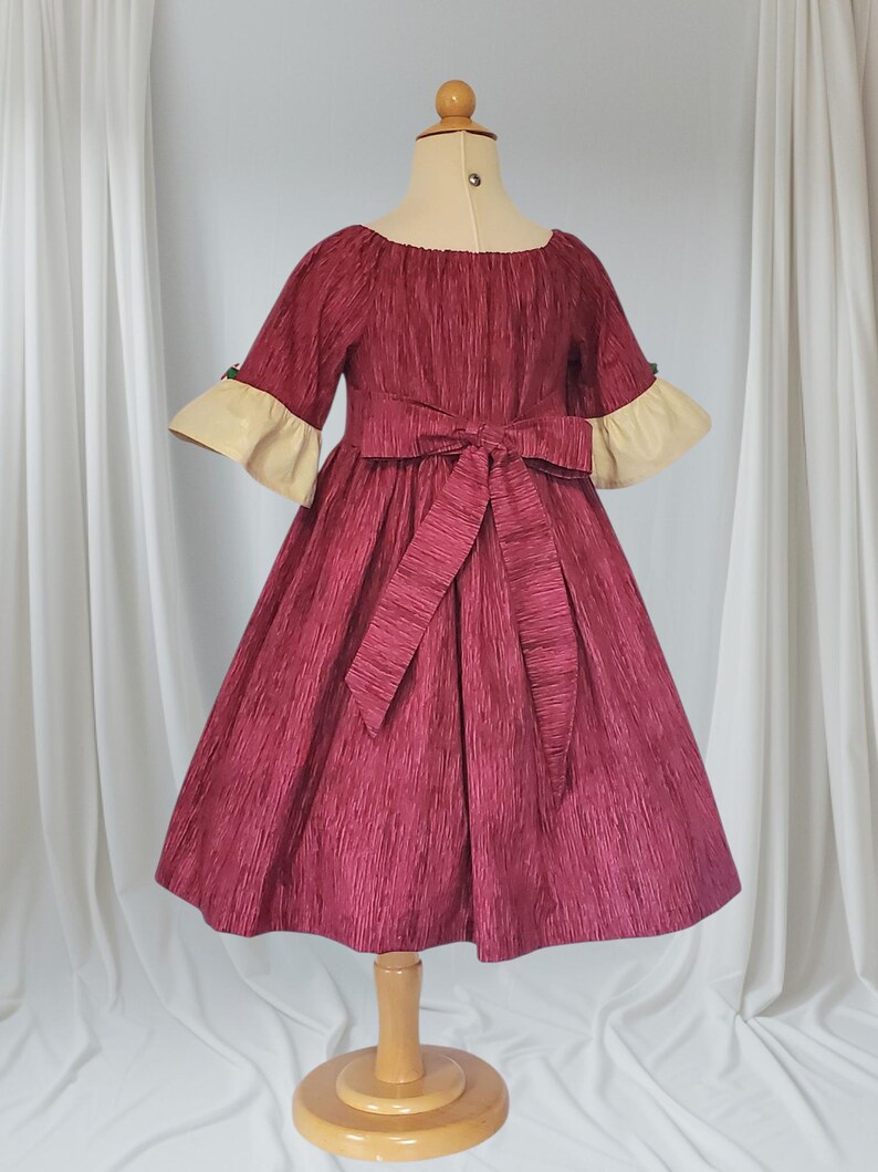 May include: A burgundy dress with textured fabric, featuring a boat neckline and elbow-length sleeves with cream-coloured cuffs. A matching bow adorns the waist. The dress is displayed on a mannequin.