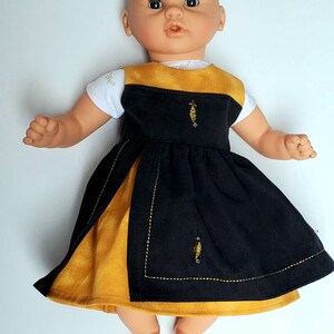 Puede incluir: Una mu&ntilde;eca con un vestido negro y amarillo mostaza con abejas bordadas. El vestido tiene una camiseta blanca con la palabra "Belle" en ella.