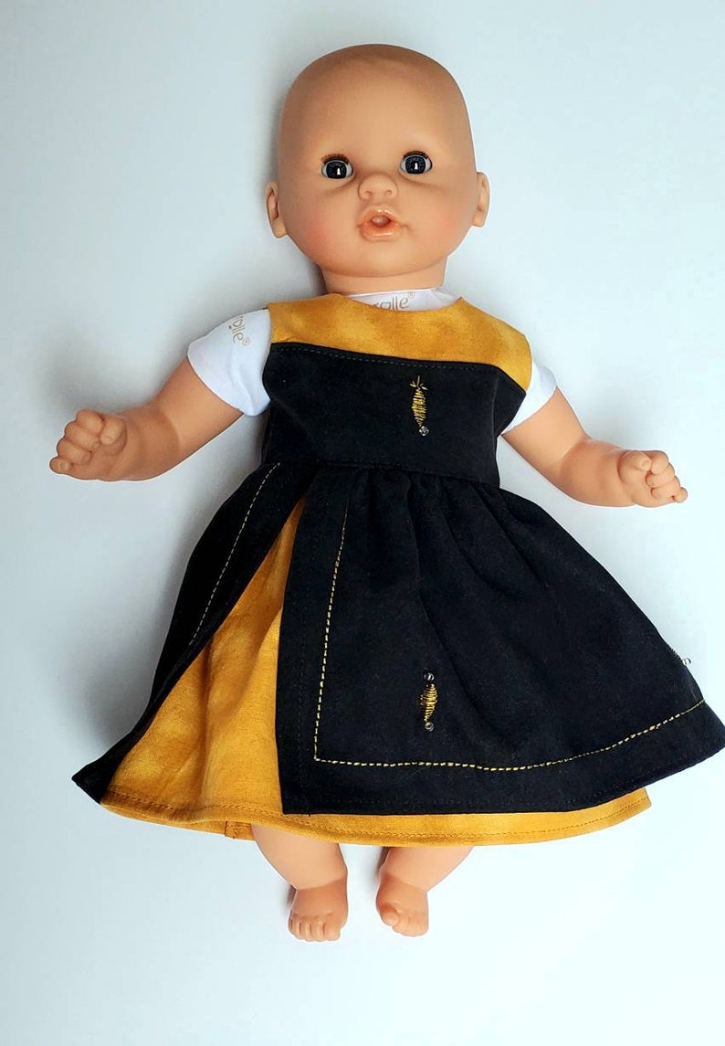 Puede incluir: Una mu&ntilde;eca con un vestido negro y amarillo mostaza con abejas bordadas. El vestido tiene una camiseta blanca con la palabra "Belle" en ella.