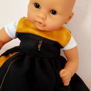 Puede incluir: Una mu&ntilde;eca beb&eacute; con un vestido negro y dorado con abejas bordadas. El vestido tiene una camiseta blanca con la palabra "walle" en ella.