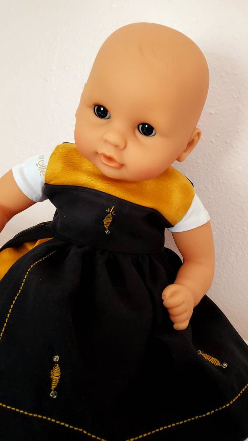 Puede incluir: Una mu&ntilde;eca beb&eacute; con un vestido negro y dorado con abejas bordadas. El vestido tiene una camiseta blanca con la palabra "walle" en ella.