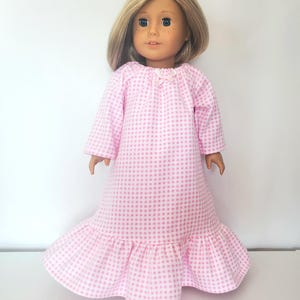 May include: A doll wearing a pink and white gingham nightgown. The long-sleeved gown has a ruffled hem and a small bow at the neckline. The doll has blonde hair and blue eyes.