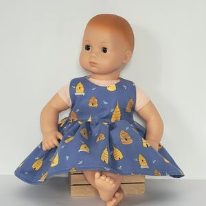 May include: A doll wearing a blue dress with a yellow beehive pattern. The doll has fair skin and red hair. The dress has short sleeves and a flared skirt. The doll is sitting on a small wooden crate.