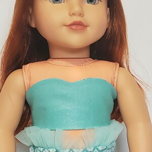May include: A doll with red hair and blue eyes wearing a mermaid costume. The costume features a teal top with a ruffled trim and a scale-patterned skirt. The doll has a fair complexion and is smiling.