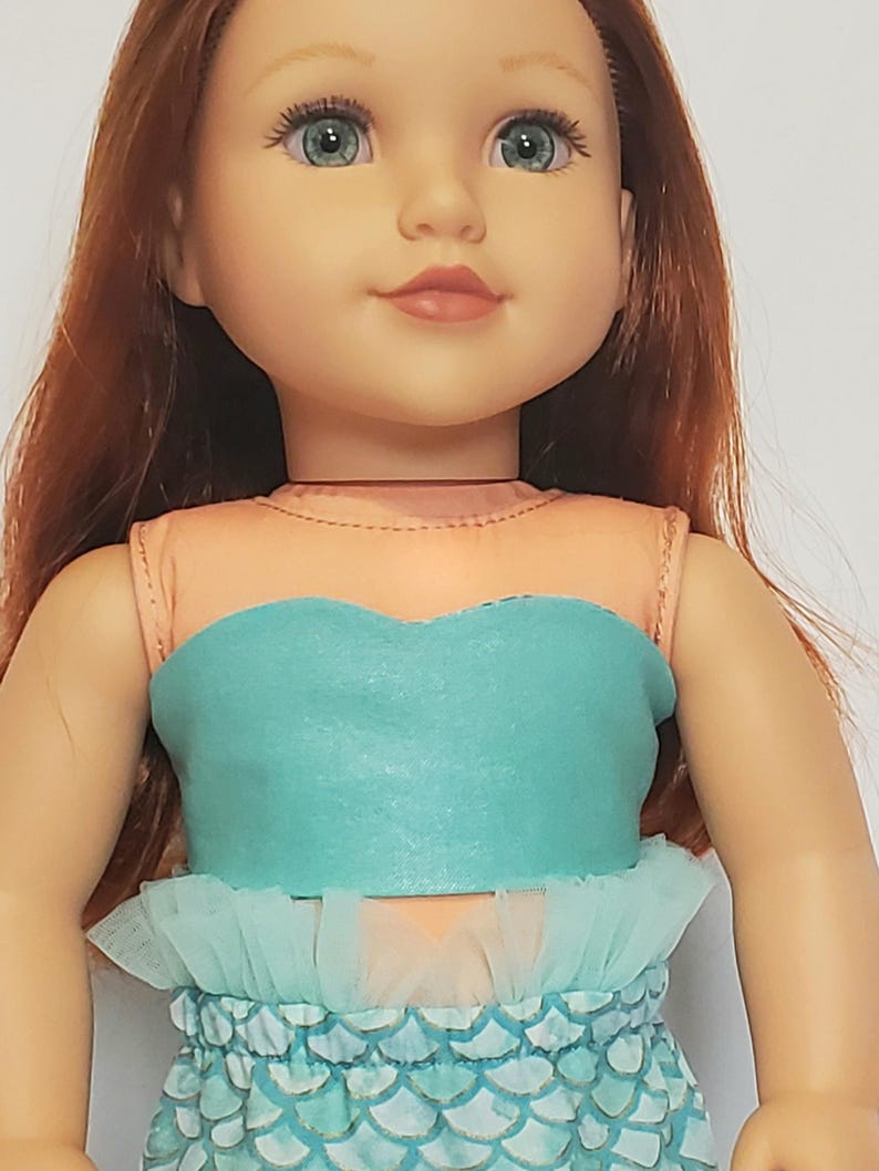 May include: A doll with red hair and blue eyes wearing a mermaid costume. The costume features a teal top with a ruffled trim and a scale-patterned skirt. The doll has a fair complexion and is smiling.