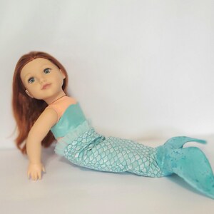 May include: A mermaid doll with red hair and blue eyes. The doll wears a teal and peach top and a teal scale-patterned tail. The doll is lying down on a white surface.