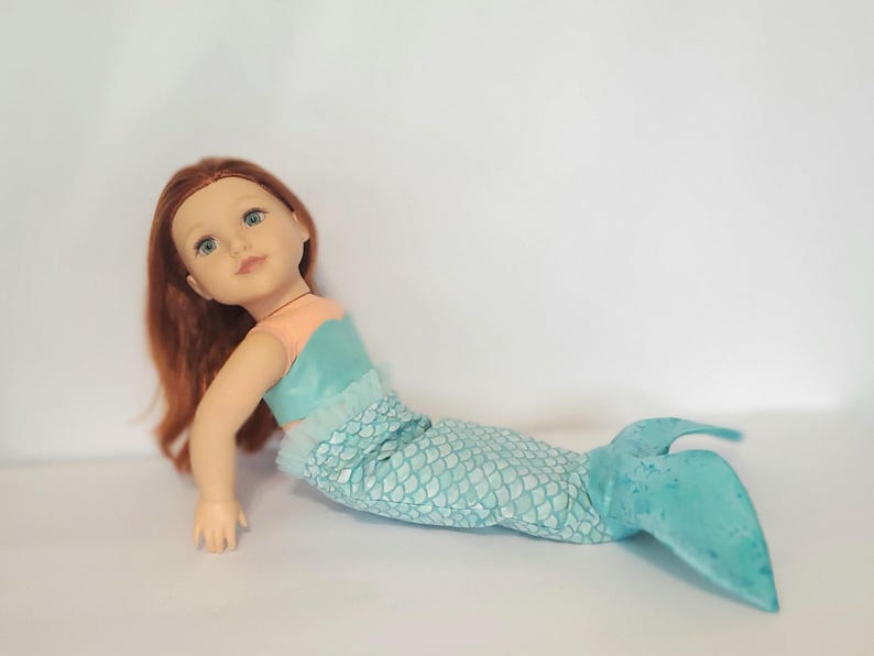May include: A mermaid doll with red hair and blue eyes. The doll wears a teal and peach top and a teal scale-patterned tail. The doll is lying down on a white surface.