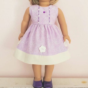 May include: A lavender doll dress with a white hem and flower accents. The sleeveless dress features a floral pattern and is adorned with white flower-shaped appliques. The doll is wearing matching purple glitter shoes.