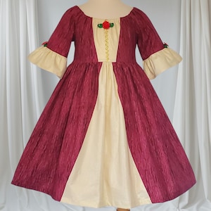 May include: A burgundy and cream-coloured dress with a vintage design. The dress features a scoop neckline, three-quarter sleeves with cream-coloured cuffs, and a full skirt. A gold trim and a red rose accent the bodice. The dress is displayed on a mannequin.