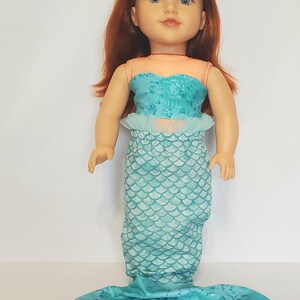 May include: A doll dressed as a mermaid. The doll has red hair and blue eyes. The mermaid outfit includes a teal top, a skirt with a scale pattern, and a teal tail. The top is a light teal color with a ruffled trim.