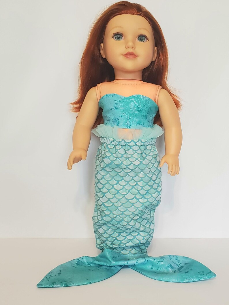May include: A doll dressed as a mermaid. The doll has red hair and blue eyes. The mermaid outfit includes a teal top, a skirt with a scale pattern, and a teal tail. The top is a light teal color with a ruffled trim.