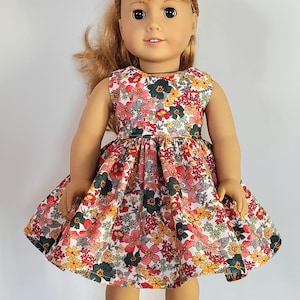 May include: A doll wearing a sleeveless floral dress with a white background and red, orange, yellow, green, and grey flowers. The doll has long, wavy, light brown hair and brown eyes. The doll is wearing white socks with lace trim and brown shoes.