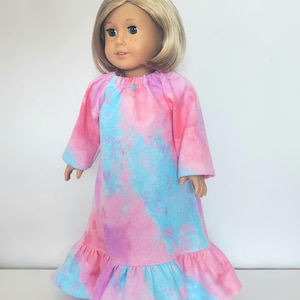 May include: A doll wearing a tie-dye nightgown in shades of pink, blue, and purple. The long-sleeved gown has a ruffled hem and a button closure at the neckline. The doll has blonde hair and is standing against a white background.