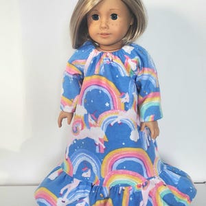 May include: A doll wearing a long-sleeved blue dress with a rainbow and unicorn pattern. The dress has a ruffled hem and a round neckline. The doll has blonde hair and is standing against a white background.