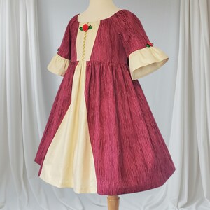 May include: A burgundy and cream-coloured dress on a mannequin. The dress has a textured fabric, a cream-coloured panel down the front, and cream-coloured bell sleeves. A red rose and gold trim adorns the bodice. The dress is designed for a child.