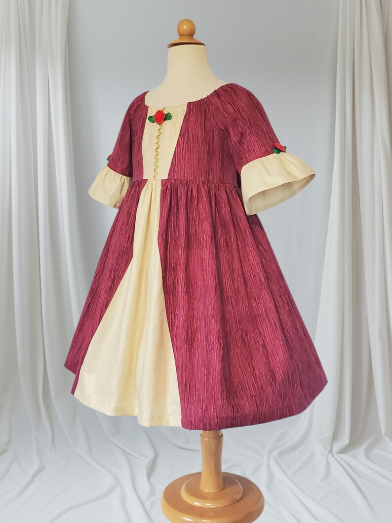May include: A burgundy and cream-coloured dress on a mannequin. The dress has a textured fabric, a cream-coloured panel down the front, and cream-coloured bell sleeves. A red rose and gold trim adorns the bodice. The dress is designed for a child.