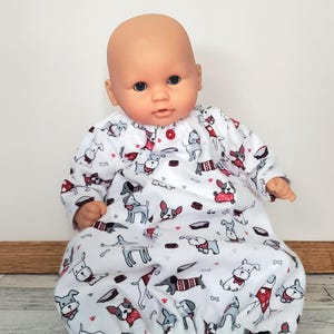 May include: A baby doll wearing a white sleep sack with a repeating pattern of dogs in various poses and red accents. The doll has a bald head and is sitting upright. The sleep sack has long sleeves and a button closure.