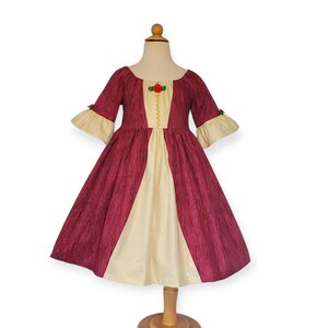 May include: A burgundy and cream-coloured dress with a vintage design. The dress features a scoop neckline, elbow-length sleeves with cream-coloured cuffs, and a full skirt. A small red rose and gold trim adorn the bodice. The dress is displayed on a mannequin.
