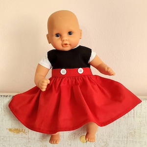 May include: A doll wearing a red skirt with a black top and white sleeves. The skirt is a vibrant red, and the top is black with white sleeves. Two white buttons adorn the waistline. The doll has blue eyes and a neutral expression.